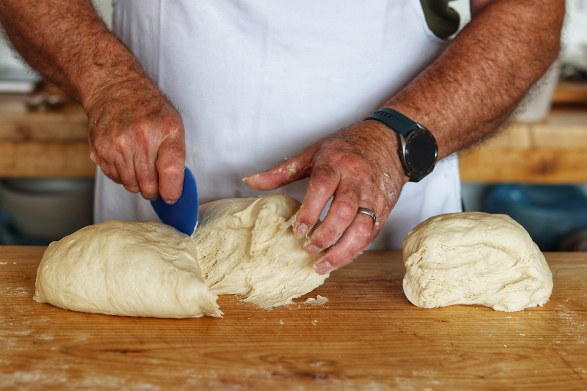 A baker in a white shirt holding a freshly shaped round of dough, candid and lit by natural kitchen light. Stand-in image — replace with a real photo of Clara before launch.
