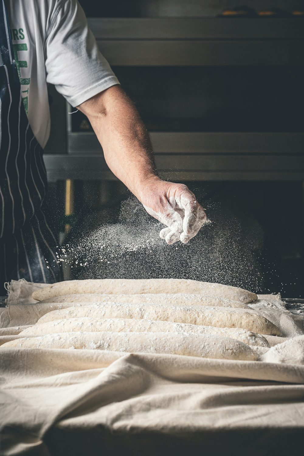 A baker in an apron sprinkling flour across a wooden work table, photographed from a low angle in warm natural light. Stand-in image — replace with a real photo of Clara.