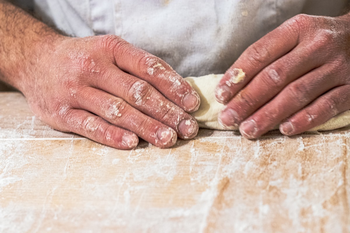 A close, detail shot of hands pressing and shaping dough on a floured wooden work surface, in warm light